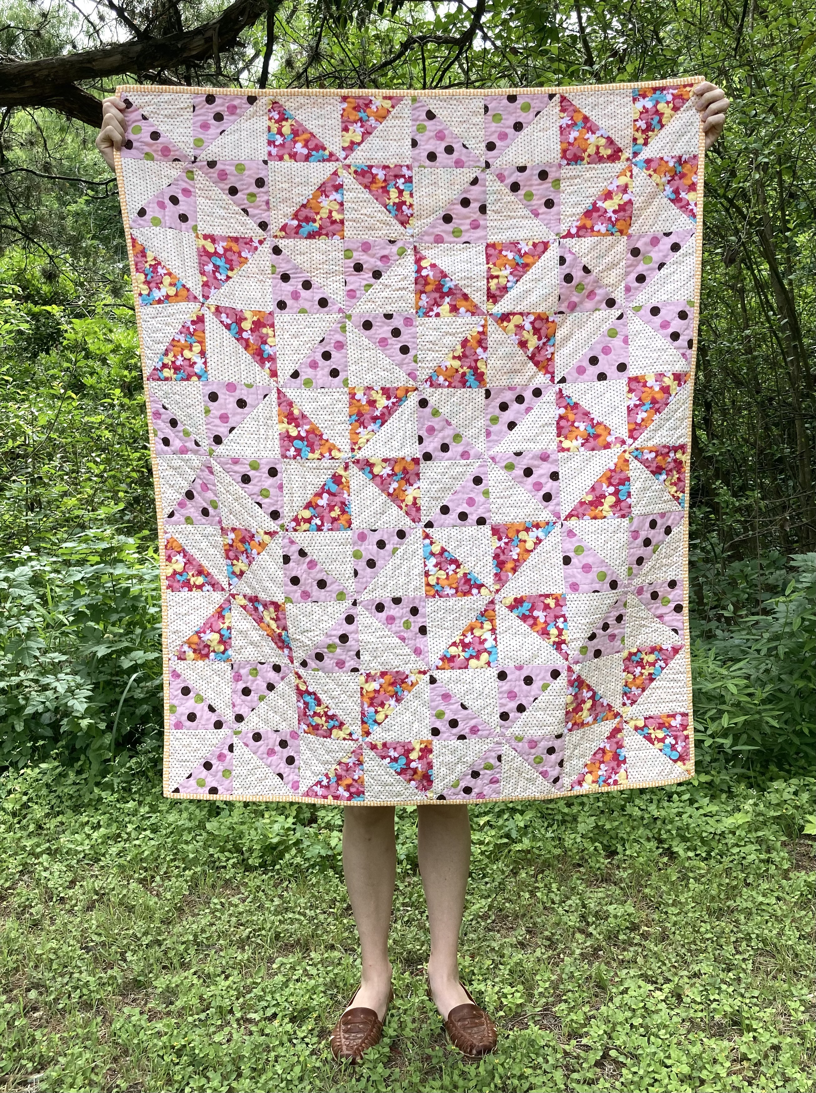 medium sized quilt with patterned pink pinwheel pattern on cream background being held by a white woman with curly hair in front of a white wall outdoors